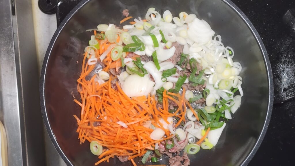 Stir-frying the partially cooked beef with onion, Korean green onion, and carrot until the vegetables soften slightly.