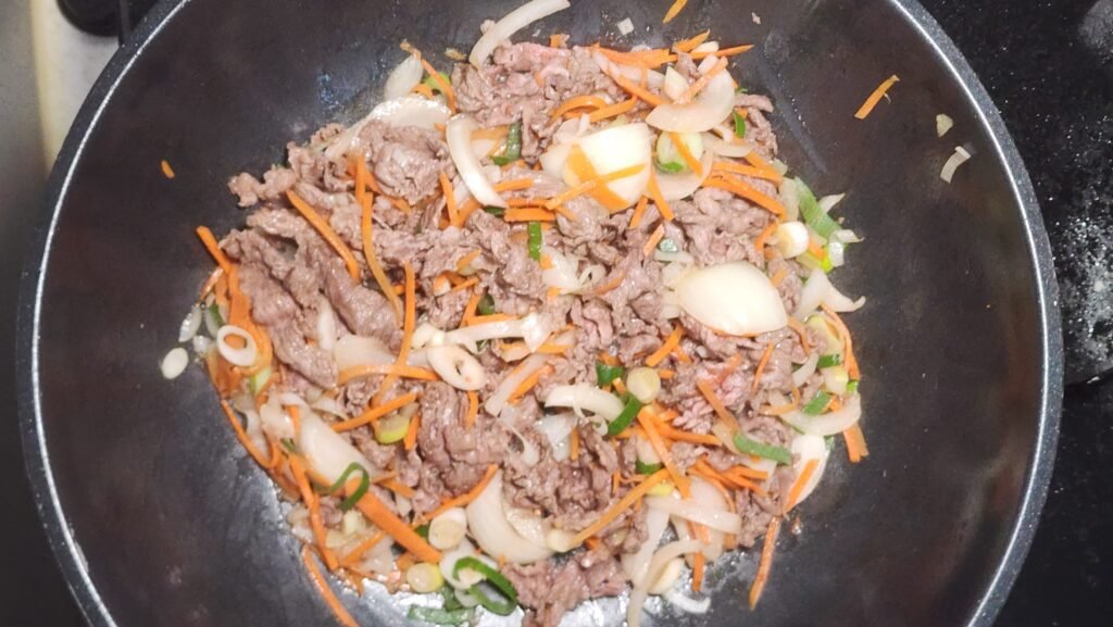Stir-frying the partially cooked beef with onion, Korean green onion, and carrot until the vegetables soften slightly.
